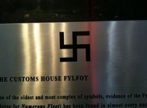 The image shows a metallic plaque with a black fylfot (a type of swastika) symbol prominently displayed in the center. Below the symbol, there is text that reads "THE CUSTOMS HOUSE FYLFOT" followed by additional text that is partially visible. The plaque appears to be part of an exhibit or informational display.