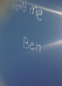 The image shows a clear blue sky with the words "marry me Ben" written in skywriting. The text appears to be created by an airplane using smoke. Sunlight is also visible, creating a lens flare effect in the image.