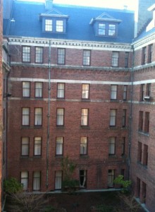 The image shows the interior courtyard of a multi-story brick building. The building has several rows of windows, some of which are lit from the inside. The courtyard is surrounded by the building on all sides, and there are a few small trees and shrubs at the ground level. The architecture appears to be older, with a traditional design and a dark roof.