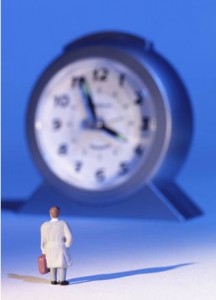 The image shows a small figurine of a person in a white coat holding a briefcase, standing in front of a large clock. The clock is significantly larger than the figurine, creating a surreal and exaggerated perspective. The background is blue, and the scene is well-lit, casting shadows on the ground.