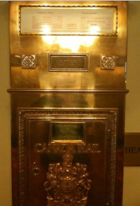 The image shows a vintage, ornate Canada Post mailbox. The mailbox is made of polished brass or a similar metal, giving it a shiny, reflective surface. It features intricate detailing, including a coat of arms and the words "Canada Mail" engraved on the front. There is a slot for mail at the top and a larger compartment below, likely for collecting the deposited mail. The overall design is elegant and historic, suggesting it might be located in a significant or older building.