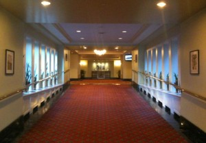 The image shows a long, well-lit hallway with a red carpet. The walls are light-colored and adorned with framed pictures. There are handrails on both sides of the hallway, and windows with plants on the left side. At the end of the hallway, there is a chandelier hanging from the ceiling and a small table or counter with decorative items. The overall ambiance is elegant and inviting.