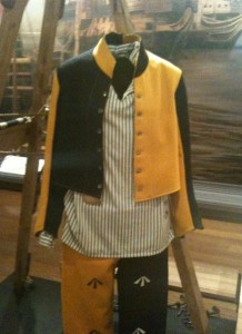 The image shows a historical costume displayed on a mannequin. The outfit consists of a yellow and black jacket with buttons, a striped shirt underneath, and matching yellow and black pants with decorative patterns. The background appears to be a museum or exhibit setting with wooden floors and a painting or mural on the wall.