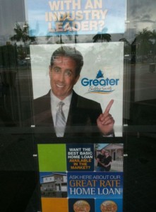 The image is a promotional poster for a home loan service. The top part of the poster features a man in a suit pointing upwards with the text "WITH AN INDUSTRY LEADER?" above him. Below the man, there is a logo that reads "Greater" with a stylized tree. The bottom part of the poster includes text that says "WANT THE BEST BASIC HOME LOAN AVAILABLE IN THE MARKET? ASK HERE ABOUT OUR GREAT RATE HOME LOAN." There are also images of houses and a family, as well as some circular icons at the very bottom. The poster is displayed on a glass surface, and there are reflections of the outside environment visible.