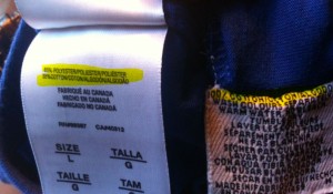 The image shows two clothing tags. The tag on the left provides information about the fabric composition, manufacturing location, and size. It states that the item is made of 100% polyester, made in Canada, and is size large (L). The tag on the right contains care instructions in multiple languages, advising on how to wash and care for the garment. Both tags are attached to a piece of blue fabric.