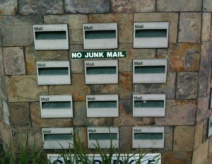 The image shows a stone wall with multiple mail slots labeled "Mail." In the center of the wall, there is a sign that reads "NO JUNK MAIL." There are some green plants at the bottom of the image.