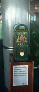 The image shows a green post box with the label "NSW MAIL" and a coat of arms. The post box is mounted on a wooden stand. A sign on the stand reads, "The Highest Working Post Box in the Southern Hemisphere." The background includes a large metal column and some greenery.