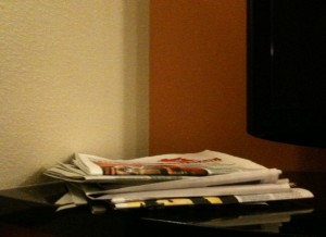 A stack of newspapers and magazines is placed on a dark surface next to a wall. The background includes a corner where two walls meet, one painted white and the other a warm orange color. Part of a television screen is visible on the right side of the image.