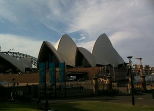 The image shows the Sydney Opera House, a famous architectural landmark in Sydney, Australia. The building features its distinctive sail-like design against a partly cloudy sky. In the foreground, there is a fenced area with some banners and a few street lamps.