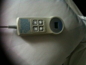 The image shows a close-up of a wired remote control device, likely for adjusting a hospital bed or a recliner. The remote has four directional buttons arranged in a square pattern and a small display screen. The device is resting on a white surface, possibly a bed or a pillow.