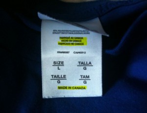The image shows a clothing label attached to a dark blue garment. The label contains information in multiple languages, including English, Spanish, and French. It indicates that the garment is made in Canada and is a size large (L). The text "MADE IN CANADA" is highlighted in yellow at the bottom of the label.