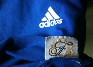 The image shows a close-up of a blue fabric with two logos. The top logo is the Adidas logo, which consists of three white diagonal stripes and the word "adidas" below it. The bottom logo is a decorative emblem with the letters "F" and "F" intertwined, surrounded by a gold wreath-like design.