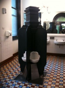 The image shows a public restroom with a unique, vintage-style urinal setup. The urinals are arranged around a central column with dark, ornate dividers providing privacy. The restroom has a tiled floor with a geometric pattern, white tiled walls, and a large window. In the background, there are two sinks with mirrors above them. The overall design of the restroom appears to be old-fashioned and decorative.