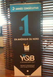 The image shows a vertical banner with text in French. The banner reads "2e ANNÉE CONSÉCUTIVE" at the top, followed by a large "#1" in the center. Below that, it says "EN AMÉRIQUE DU NORD" and features a logo with stars and the text "ASQ 2010 AWARD WINNER." At the bottom, it has the logo "YQB" with the tagline "LE PASSAGER AVANT TOUT" and mentions "Aéroport International Jean-Lesage de Québec." The banner is placed in front of a wooden slatted background.