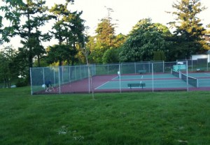 The image shows a fenced tennis court situated in a park-like area with green grass and trees. The court is empty, and there is a bench inside the fenced area. The surrounding area is lush with greenery, and the sky appears to be clear, indicating a pleasant day.