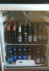 The image shows the inside of a refrigerator filled with various bottles of beer. The bottles are neatly arranged in rows on two shelves. The brands visible include Guinness, Heineken, Corona, and Budweiser, among others. The refrigerator door is open, and the bottles are standing upright.