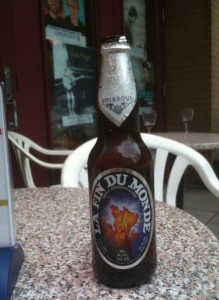 The image shows a bottle of beer labeled "La Fin Du Monde" placed on a speckled granite table. The bottle has a silver cap and a label featuring an image of a fiery map. In the background, there are white plastic chairs and a glass door with posters on it.