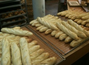 The image shows several freshly baked baguettes arranged on cooling racks in a bakery. There are multiple rows of baguettes, and in the background, there are more baked goods on shelves. The setting appears to be a bakery or a bread shop.