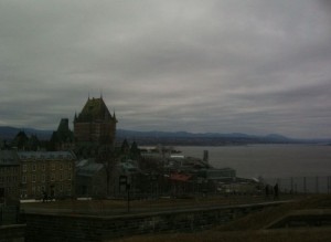 The image shows a cloudy day with a view of a large, historic building with a steep, green roof, likely a castle or a grand hotel, situated near a body of water. The building is surrounded by other smaller buildings and there are mountains visible in the background across the water. The overall atmosphere is overcast and somewhat gloomy.