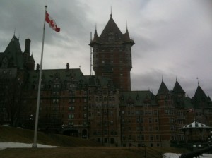 The image shows a large, historic building with a castle-like appearance, featuring multiple turrets and a central tower. The building is made of brick and has a green roof. In the foreground, there is a flagpole with a Canadian flag flying. The sky is overcast, giving the scene a somewhat gloomy atmosphere. The building is situated on a grassy area with a few patches of snow.