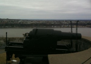 The image shows an old, large cannon mounted on a platform overlooking a body of water. In the background, there is a cityscape with numerous buildings and a cloudy sky. The cannon appears to be part of a historical site or fortification.