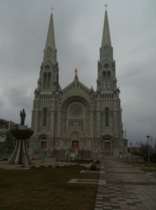 The image shows a large, ornate cathedral with two tall spires. The building is constructed in a Gothic architectural style, featuring intricate details and a large circular stained glass window above the main entrance. In front of the cathedral, there is a statue of a person standing on a pedestal. The sky is overcast, and the surrounding area includes a paved walkway and some grassy areas.