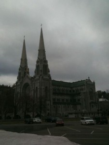 The image shows a large, historic church with two tall spires. The building is made of stone and has intricate architectural details. The sky above is overcast, and there are a few cars parked in the parking lot in front of the church. The surrounding area appears to be quiet and somewhat empty.