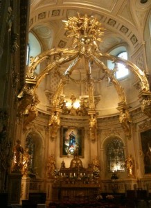 The image shows the interior of a richly decorated church or cathedral. The focal point is an ornate altar adorned with golden sculptures and intricate designs. Above the altar, there is a large, elaborate golden structure with angelic figures and decorative elements. The walls and ceiling are also intricately designed, with arches and detailed artwork. A painting or stained glass window depicting a religious figure is prominently displayed behind the altar. The overall atmosphere is grand and opulent, reflecting the architectural and artistic splendor of the space.