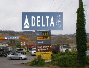 The image shows a Delta gas station with a large sign displaying the Delta logo at the top. Below the logo, the price of fuel is listed as $3.99. The sign also advertises "easy PAY" and mentions "Diesel" fuel. Additionally, there is a smaller sign that reads "Got gunky build-up?" with a reference to a product called "Supreme Diesel." The gas station is situated in a somewhat rural area with hills and trees in the background. Several vehicles are parked near the gas pumps.