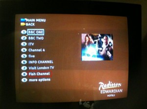 The image shows a television screen displaying a menu for selecting TV channels. The menu options listed are:

1. BBC ONE
2. BBC TWO
3. ITV
4. Channel 4
5. five
6. INFO CHANNEL
7. Visit London TV
8. Fish Channel
9. more options

The screen also has a small preview image of a TV show or movie, and the bottom right corner displays the logo for "Radisson Edwardian Hotels." The top left corner has options for "MAIN MENU" and "BACK."