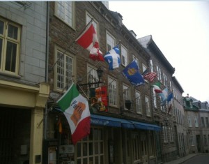 The image shows a row of buildings with multiple flags hanging from poles attached to the facade. The flags include the Canadian flag, the Quebec flag, the United States flag, the Mexican flag, and several others. The buildings have a historic architectural style, with stone walls and multiple windows. The street appears to be in an older part of a city, possibly a tourist area.