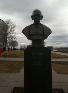 The image shows a bust statue of a person on a pedestal. The statue is located outdoors, with a cloudy sky in the background. There are trees and a few people walking in the distance. The pedestal has an inscription, but the text is not clearly readable in the image. The surrounding area appears to be a park or a campus with paved walkways and grassy areas.