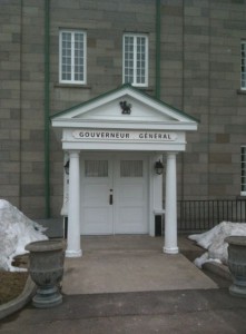 The image shows the entrance to a building with a sign above the door that reads "Gouverneur Général." The entrance features a small portico with four white columns supporting a triangular pediment. There are two white doors beneath the sign, and the building's exterior is made of gray stone. Snow is visible on the ground on either side of the entrance, and there are two large planters flanking the walkway leading to the doors.