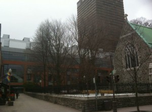 The image shows a street scene with a mix of modern and older architecture. On the right side, there is a stone building with a green roof, which appears to be a church. In the background, there is a tall, modern building. The street is lined with bare trees, indicating it might be winter or early spring. There is a sidewalk with a few people walking and a small market or shop with a colorful awning on the left side. The overall atmosphere is overcast and somewhat gloomy.