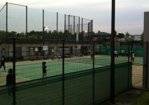 The image shows an outdoor tennis court surrounded by a high fence. Several people are on the court, some playing tennis while others are standing or walking around. The court is lit by overhead lights, and there are buildings and trees in the background. The sky appears to be overcast or during dusk.