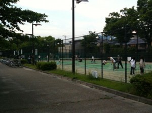 The image shows an outdoor tennis court surrounded by a tall fence. Several people are playing tennis on the court, while others are standing or walking nearby. The area is lined with trees and there is a paved path running alongside the court. Bicycles are parked along the fence on the left side of the image. The scene appears to be in a park or recreational area.