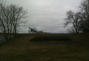 The image shows a historical site with a cannon placed on a grassy hill. The sky is overcast, and there are leafless trees on either side of the hill. At the base of the hill, there is a sign with the text "Je Me Souviens," which is French for "I Remember." The overall atmosphere is somber and reflective.