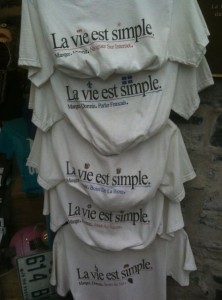 The image shows a display of five white t-shirts hanging vertically. Each t-shirt has the phrase "La vie est simple." printed on it, which translates to "Life is simple." in English. Below this phrase, each shirt has additional text in French, which varies slightly between the shirts. The t-shirts are displayed against a stone wall.