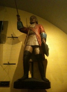 The image shows a statue of a historical figure dressed in period clothing. The figure is wearing a red coat, white shirt, and black boots, and is holding a sword in one hand and a hat in the other. The statue is mounted on a pedestal against a yellowish wall. The lighting is dim, casting shadows on the wall behind the statue.