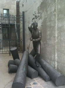 The image shows a bronze statue of a man standing on top of several cylindrical logs. The man is depicted in a working pose, holding a long pole with one hand and shielding his eyes with the other, as if looking into the distance. The statue is situated against a concrete wall, and there is a metal gate and some stairs in the background.