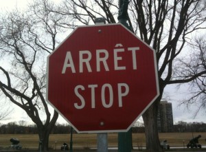 A red octagonal stop sign with the words "ARRÊT STOP" in white letters. The sign is mounted on a green pole, and there are bare trees and a park area in the background. A tall building is visible in the distance.