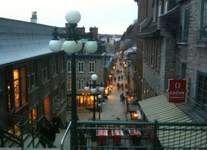 The image shows a narrow, cobblestone street in a quaint town, lined with historic buildings. The street is illuminated by street lamps and shop lights, creating a warm and inviting atmosphere. People are walking along the street, and there are various shops and cafes visible. A sign for a store or restaurant named "Cache" is prominently displayed on the right side of the image. The scene appears to be taken from an elevated position, possibly from a staircase or balcony, providing a view down the street. The overall ambiance suggests a charming, possibly European, town.