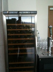 The image shows a wine cooler with a glass door, revealing several wooden shelves inside. To the right of the wine cooler, there is a countertop with a beer tap and a row of empty wine glasses. The background includes a wall and a door.