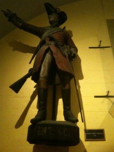 The image shows a statue of a historical figure dressed in a military uniform. The figure is wearing a tricorn hat, a long coat, and boots, and is holding a rifle in one hand while pointing forward with the other. The statue is mounted on a pedestal, and there is a plaque on the wall next to it. The lighting is dim, casting shadows on the wall behind the statue.