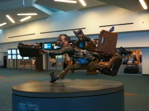 A sculpture of a man running with luggage is displayed in an airport terminal. The man appears to be in a hurry, with his body leaning forward and one leg extended behind him. He is holding a briefcase in one hand and has various bags and suitcases strapped to his back. The sculpture is mounted on a circular pedestal. The background shows the interior of the airport with seating areas and signs.