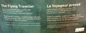 The image shows a bilingual information plaque for a sculpture titled "The Flying Traveller" (English) and "Le Voyageur pressé" (French). The sculpture was created by Patrick Amiot and Brigitte Laurent in 1996 and is made of hand-painted cast fiberglass. It was donated by Jake and Judy Kerr. The English text describes the sculpture as capturing a harried moment in the life of every traveler, while the French text conveys a similar message. The plaque also includes the YVR logo, indicating the location of the sculpture.