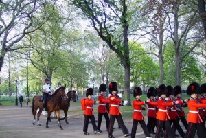 The image shows a group of soldiers in red uniforms and black bearskin hats marching in formation through a park. To the left, a mounted police officer on a horse is also visible. The park is lush with green trees and a pathway where the soldiers and the officer are moving. The scene appears to be a ceremonial or official event.