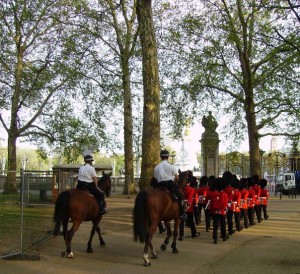 The image shows a group of guards in red uniforms and black hats marching in formation, accompanied by two mounted guards on horseback. The scene is set in a park with tall trees and a fenced area. The guards appear to be part of a ceremonial procession.