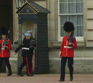 The image shows three guards in uniform outside a building. Two of the guards are wearing red coats and black bearskin hats, while the third guard, who is in the middle, is wearing a dark uniform with a light blue beret. The guard on the right is looking down at something in their hand. There is a small guardhouse in the background, and the building has large windows with white frames.