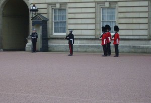 The image shows a group of guards in traditional uniforms standing in front of a historic building. Three guards are wearing red coats and black bearskin hats, while another guard in a dark uniform stands facing them. Another guard is standing near an archway to the left. The building has large windows and a stone facade. The scene appears to be a formal guard ceremony or changing of the guard.